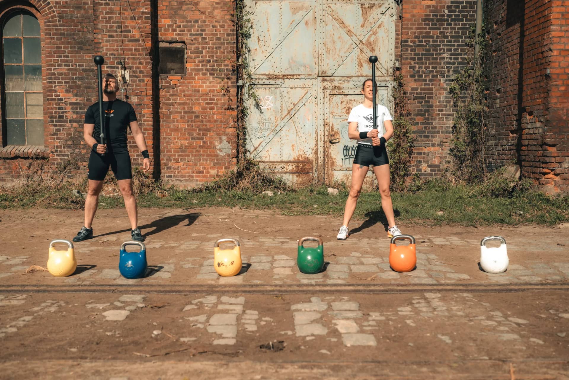 Incore Sports athletes with macebells and a row of competition kettlebells at an industrial yard