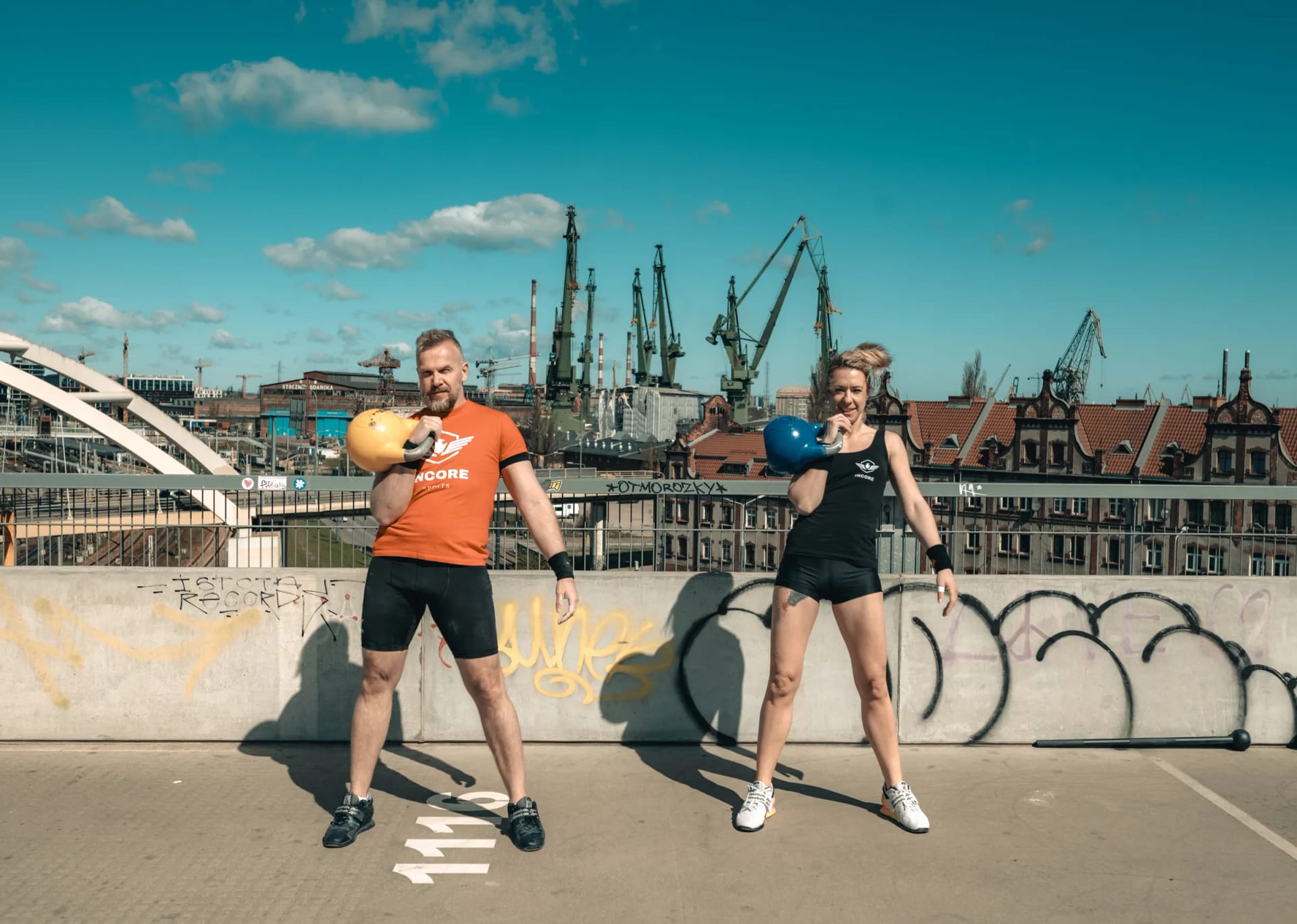 Two Incore Sports athletes holding competition kettlebells on a rooftop with shipyard cranes behind them